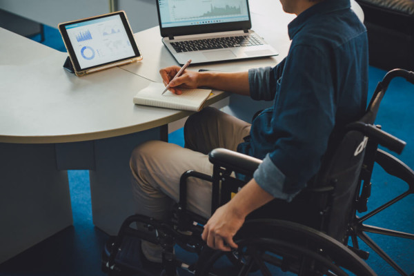 Person im Rollstuhl arbeitet an Tisch mit Block Laptop und Tablet